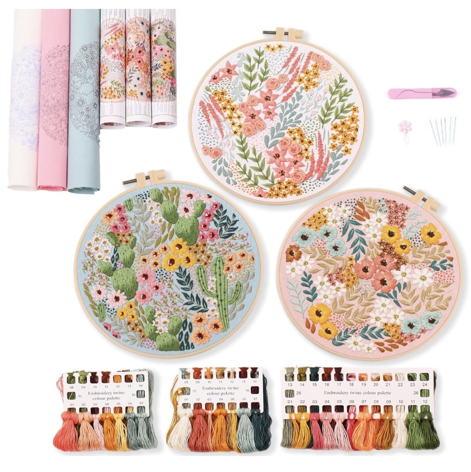 Flat lay of colorful embroidery supplies: three floral and cactus embroidered hoops, four rolled fabric patterns, a pink seam ripper, six needles, and three floss cards with various colored threads—perfect for trying out my favorite flower crafts.